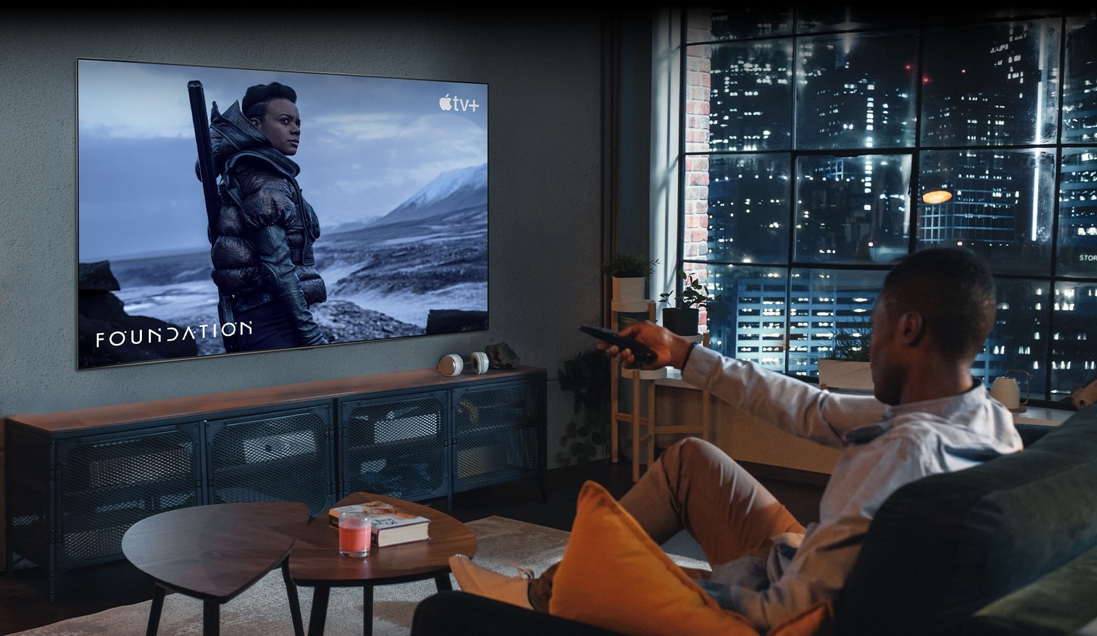 Um homem está sentado no sofá e a ver TV. O homem está com um controlo remoto na mão e na TV está uma imagem de uma cena de 'Foundation' da Apple TV+.